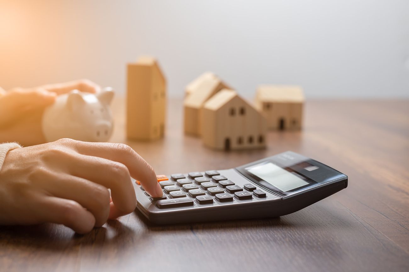 The hand is pressing calculators, piggy bank with wooden house. buy or rent question on note with calculators on desk. 