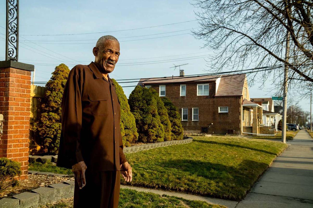 A man wearing brown, standing in front of his house