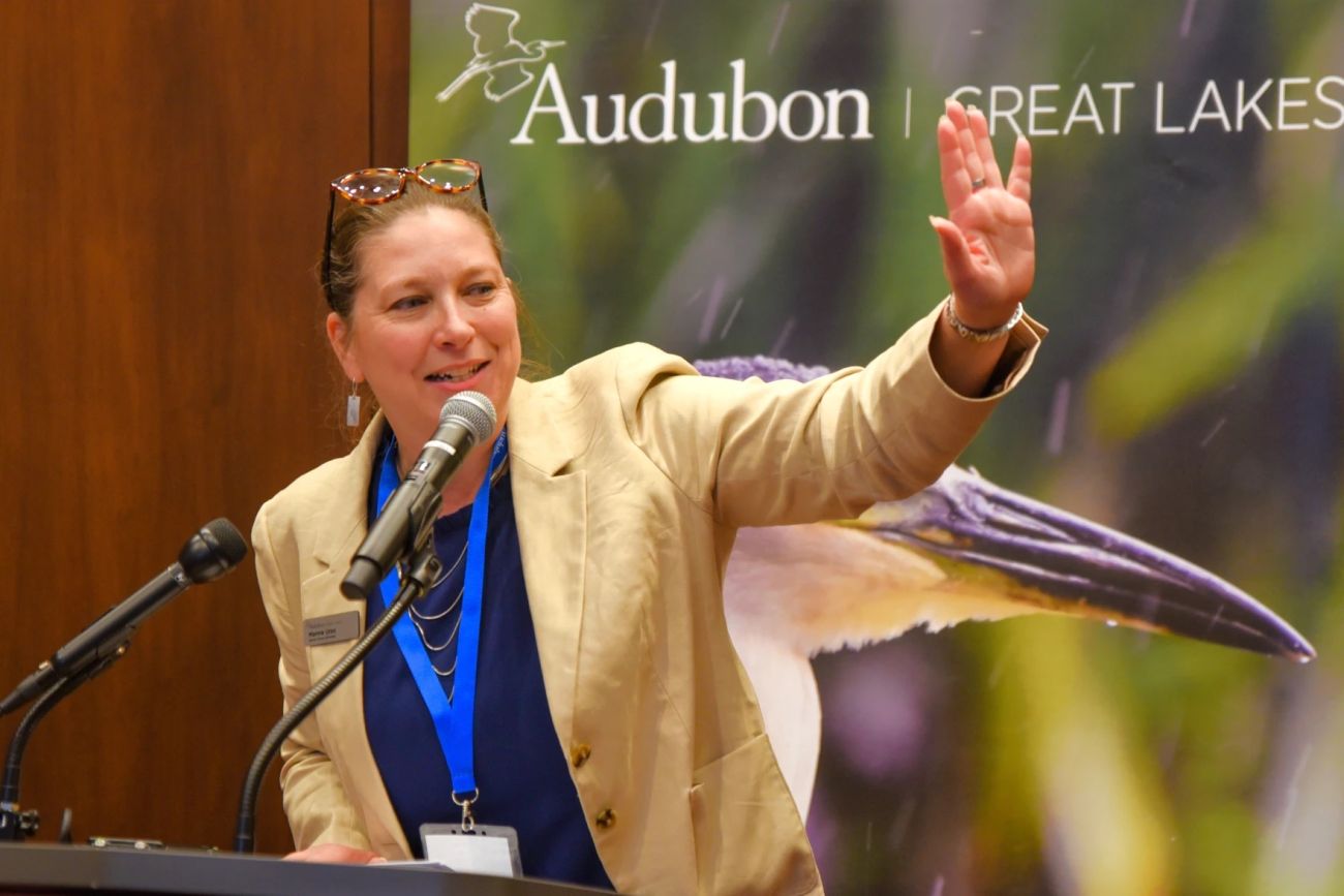 Marnie Urso is wearing a tan jacket and blue shirt. She is speaking into a microphone. There is a picture of the bird in the background