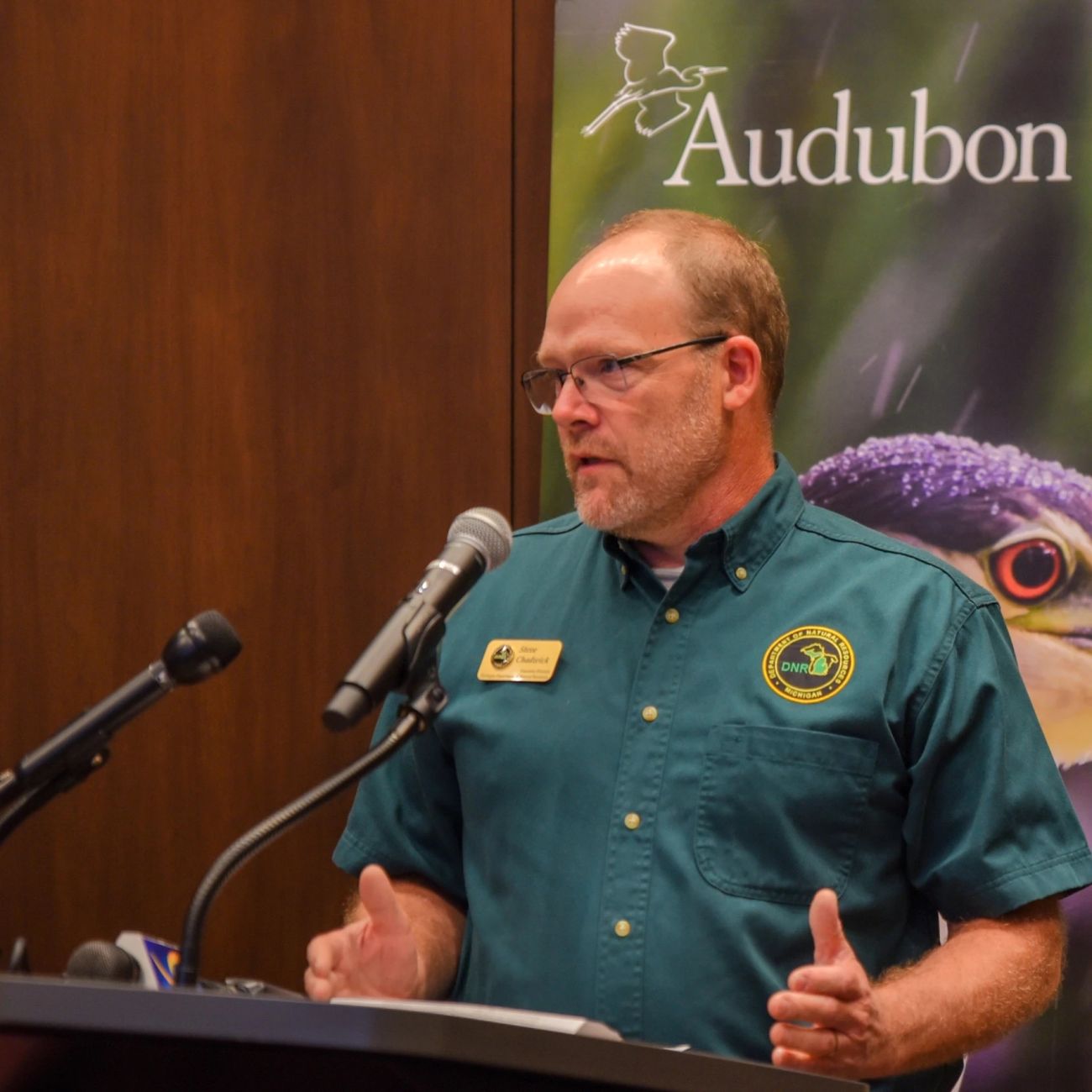 Steve Chadwick wears a green DNR shirt. He is speaking in a microphone. A picture of a bird is in the background