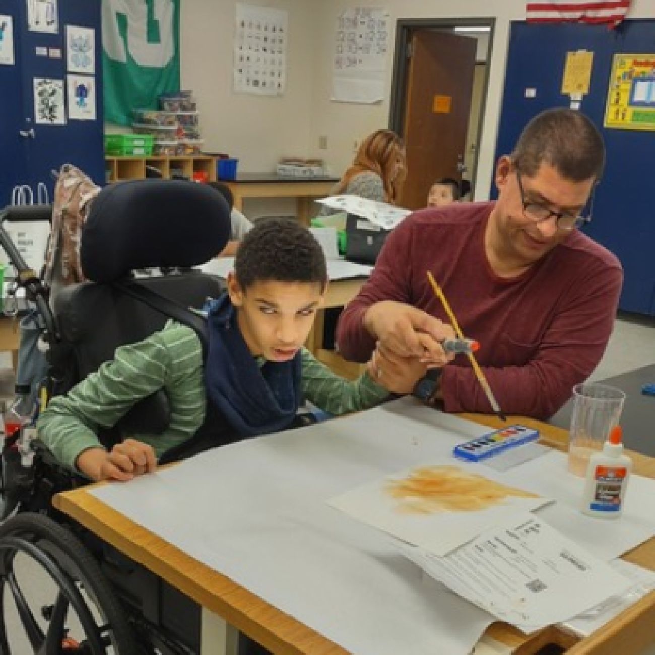 Child in a wheelchair sits at a desk with assistance