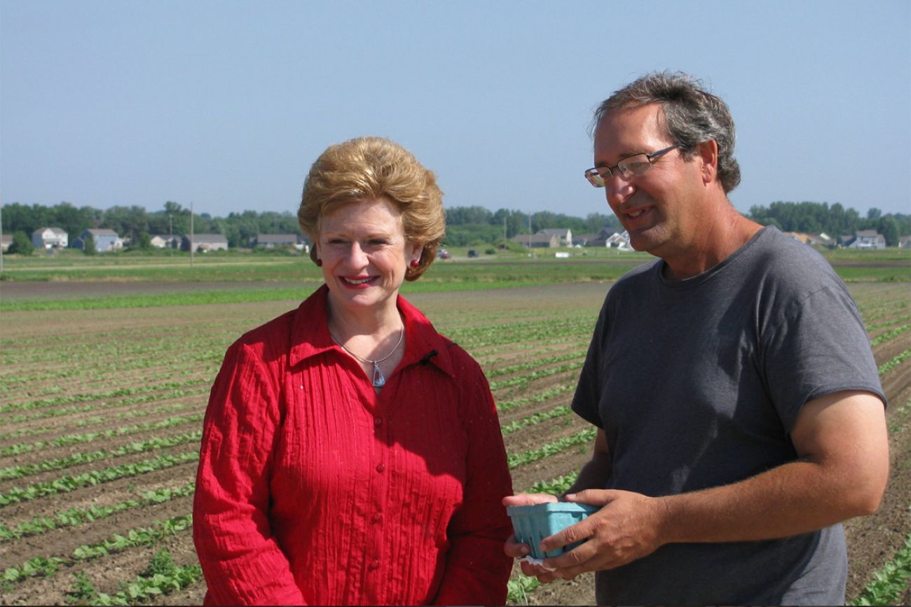 Debbie Stabenow is wearing red. Roger Victory is wearing a grey t-shirt. They are at a farm