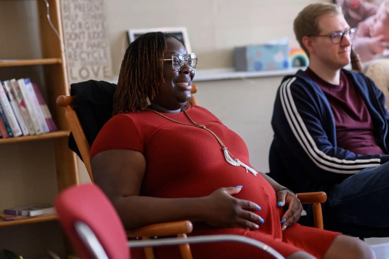 Asia LoveLady sits in a chair. She is pregnant and wearing a red dress