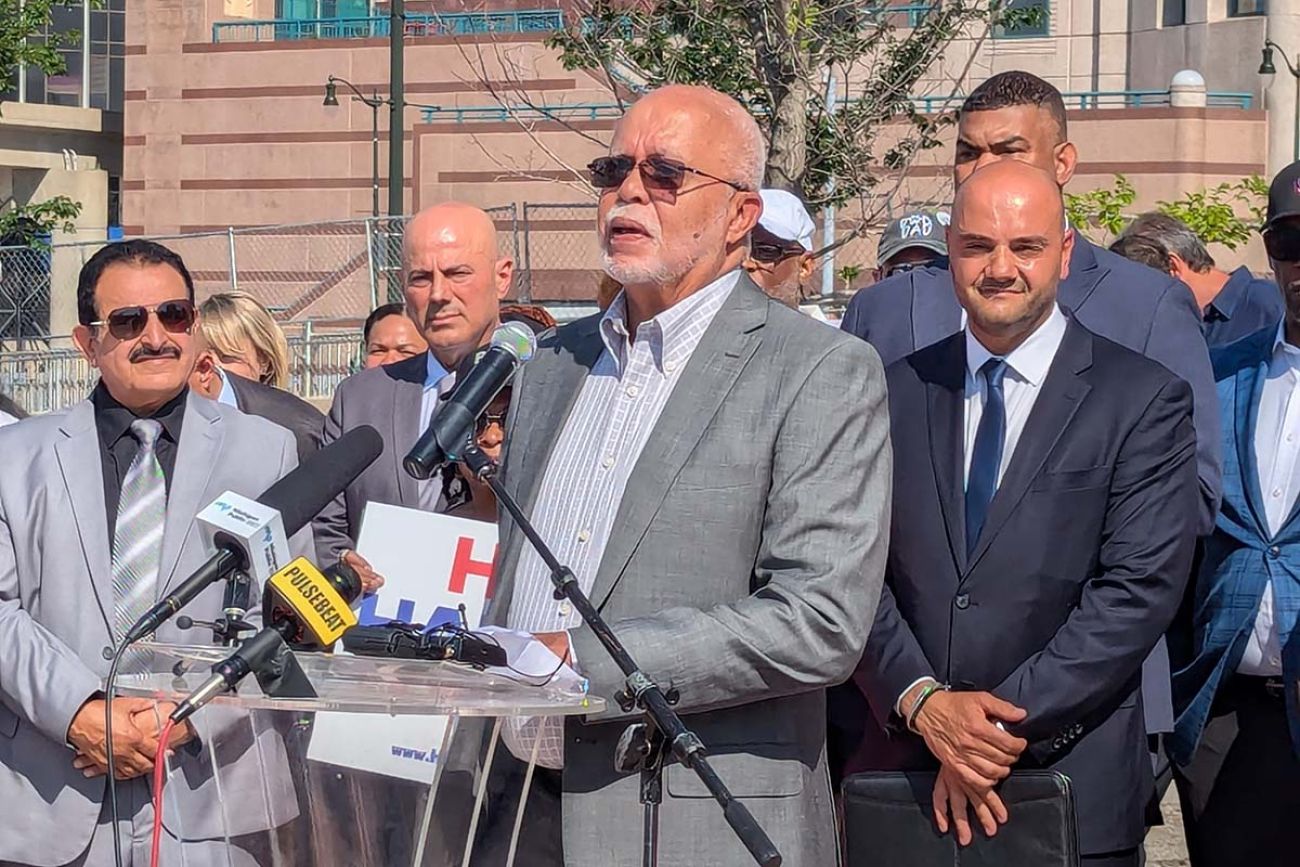 Warren Evans, wearing a grey jacket, speaks into a microphone. People are standing behind him