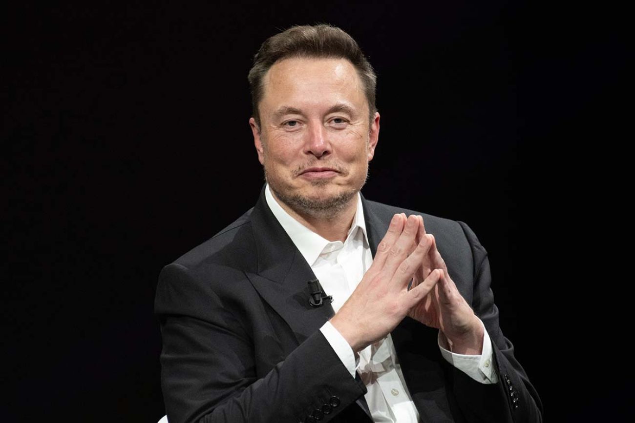 Elon Musk in a black suit, sitting in front of black background