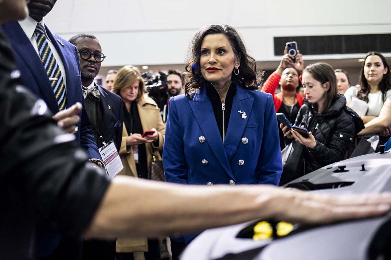 Gov. Gretchen Whitmer on the floor of the Detroit Auto Show. People at the show are around her. Whitmer is standing in front of a car