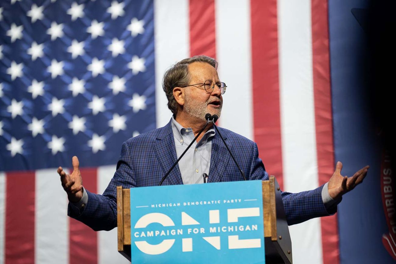 Gary Peters speaking into a microphone, an American flag behind him