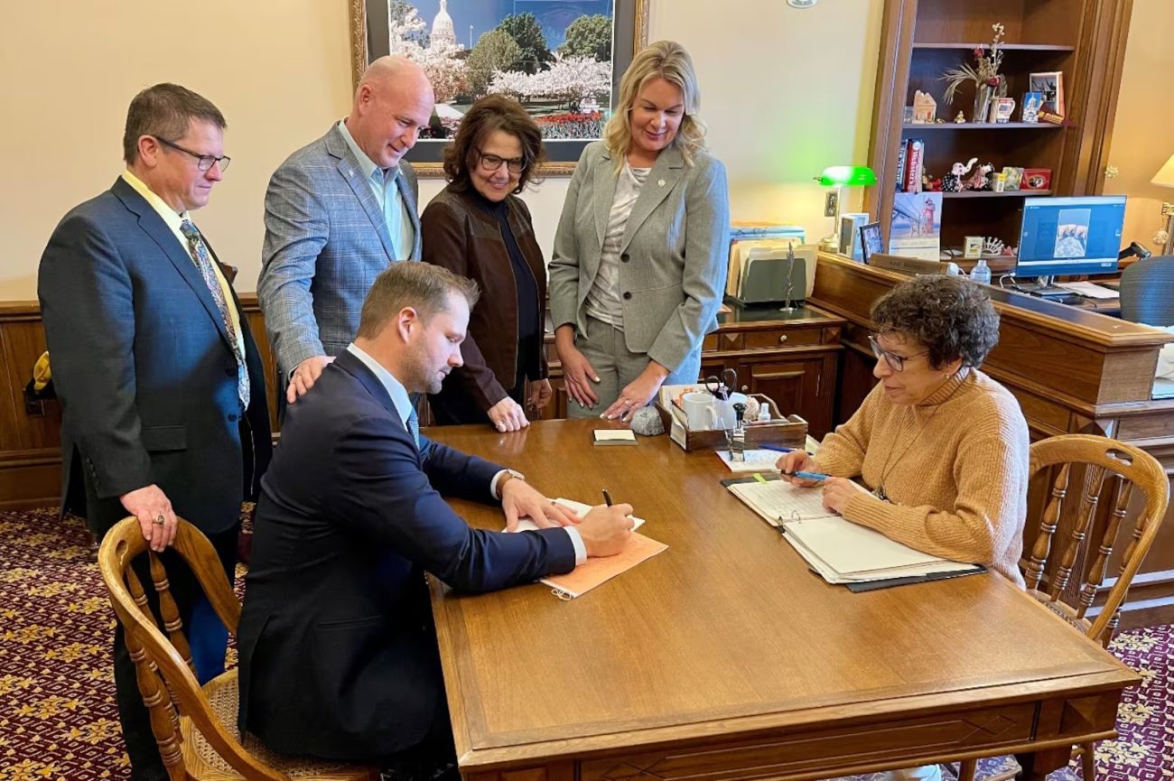 Many Michigan lawmakers sit and stand around a table. 