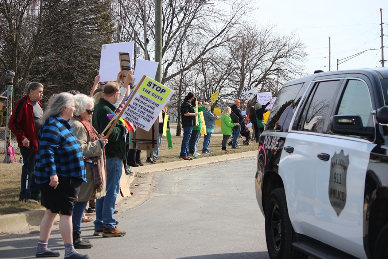 Many people on the side with the road with signs. One says "Stop the Cuts" along with several items.