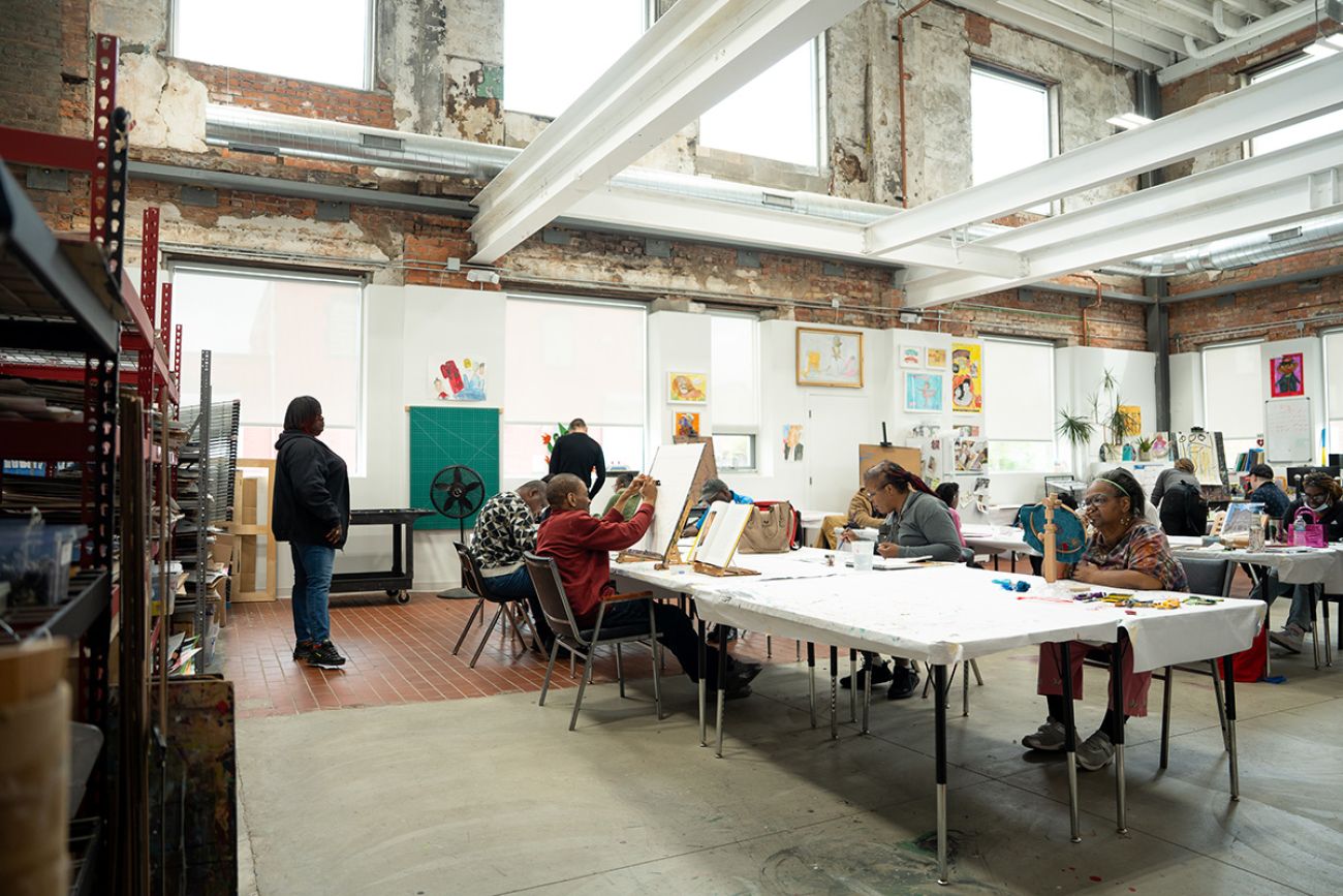 Many people are sitting at long tables while they work on their artwork.