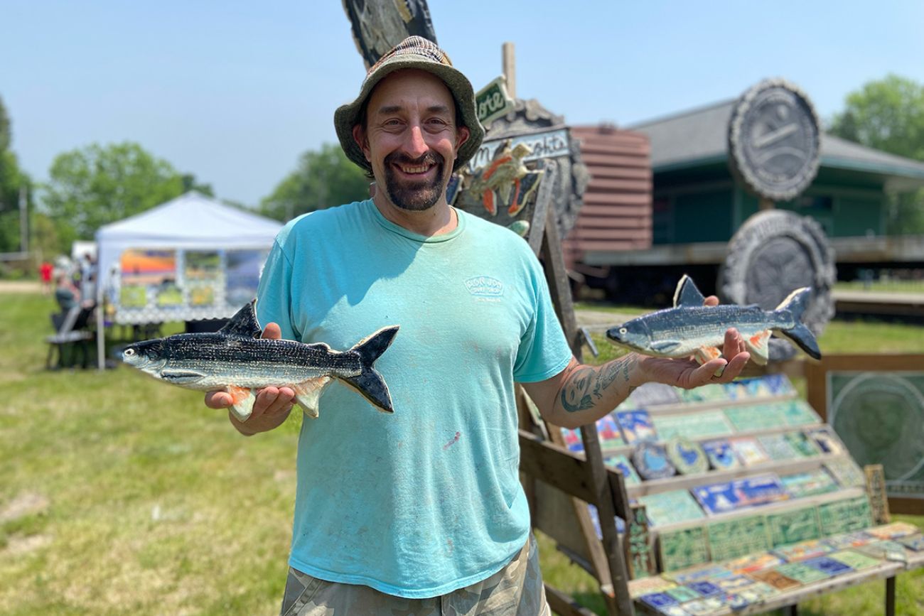 Leif Sporck holds two handcrafted tiles shaped like fish. He is outside and wearing a blue t-shirt.