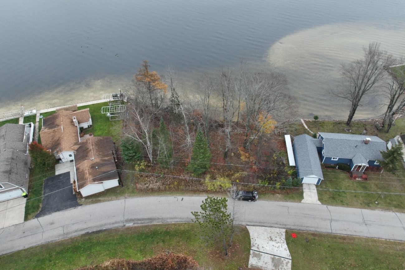 A plot of land filled with trees is seen between two properties with houses on them and a lake beyond