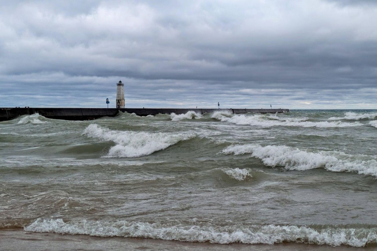 A lake is seen with big waves flowing toward shore and a lighthouse at the end of a pier in the background