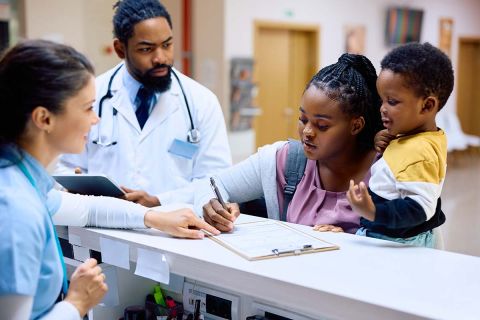 African American mother holding her small son while filling medical paperwork at reception desk in the hospital.