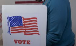 man in blue sweater and face hidden, casting vote in booth at polling station. US flag on wall in background