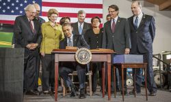 Then-President Barack Obama sitting behind a table. He is surrounded by several lawmakers. An American flag is behind the lawmakers