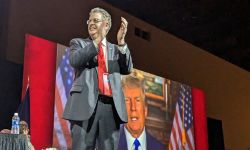 Matthew DePerno standing on a stage. A video of Donald Trump is behind him