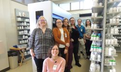 Pharmacy employees pose in front of prescription-filling machine