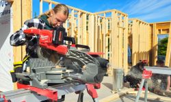 A high school-aged girl works a saw in the foreground while others work in the background of a home under construction